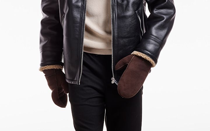 Person som har på seg brune votter med pelskant, svart skinnjakke og beige genser mot hvit bakgrunn.