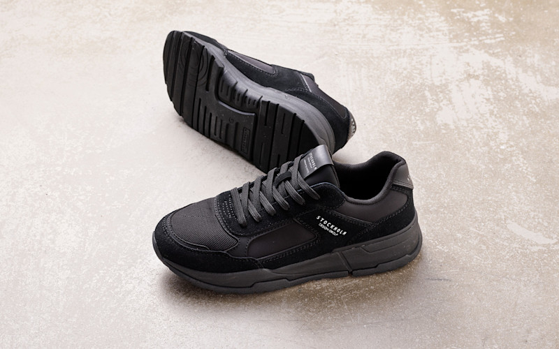 Svart sneaker med teksturen "Stockholm Design Group" p å siden, plassert på en lys betonggulv.