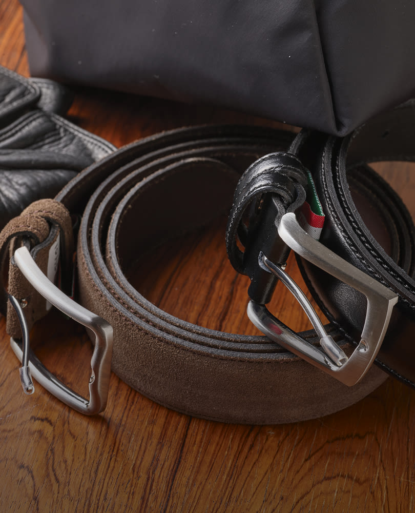 To svarte og brune skinnbelter fra Stockholm Design Group med metallspenner ligger sammenrullet på et tregulv, ved siden av et svart stoff.