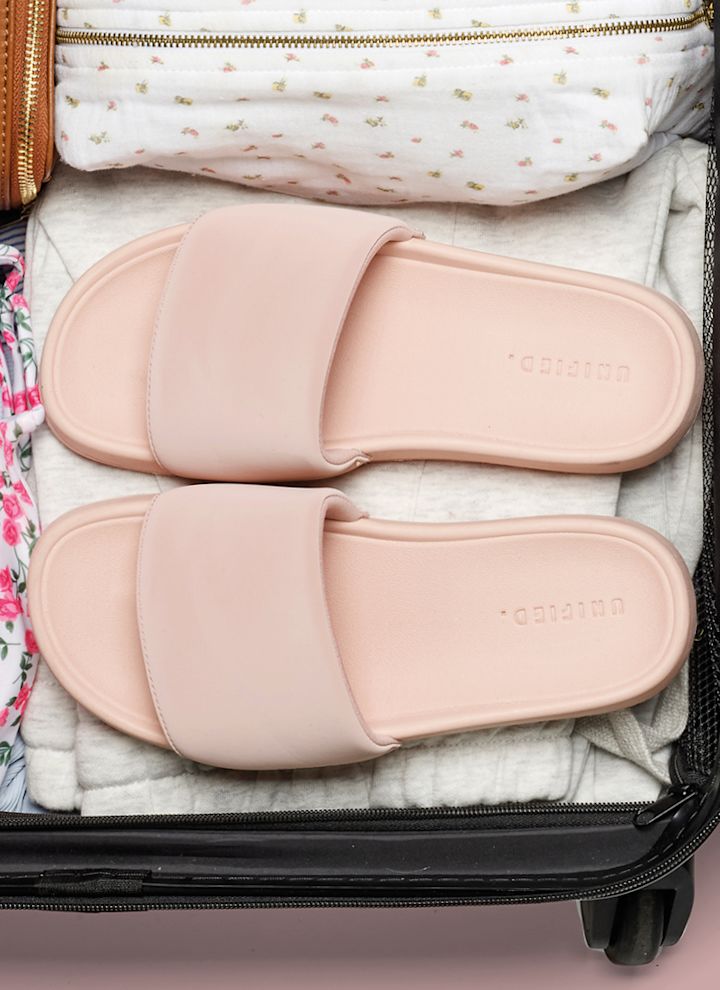 Lyserøde, minimalistiske slippers plassert i en åpen koffert pakket med klær og en brun veske.