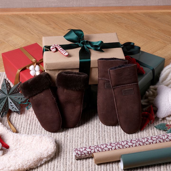 Bruna mockasiner med fluffig pälskant framför julklappar inslagna med gröna och röda band på en stickad matta.
