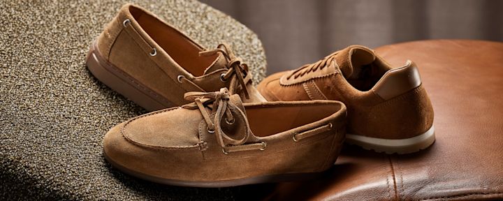 Brune semsket skinnsko - en båtsko og en casual sneaker - plassert på teppe og læroverflate i varm belysning.