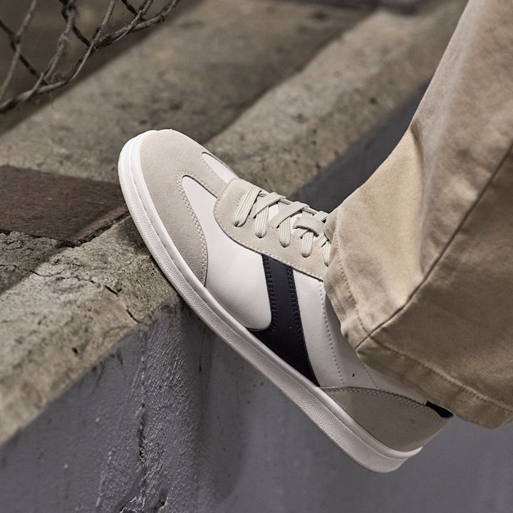 Kremhvit sneaker med mørkeblå detaljer og beige snøring, brukt med lyse bukser på betongunderlag.