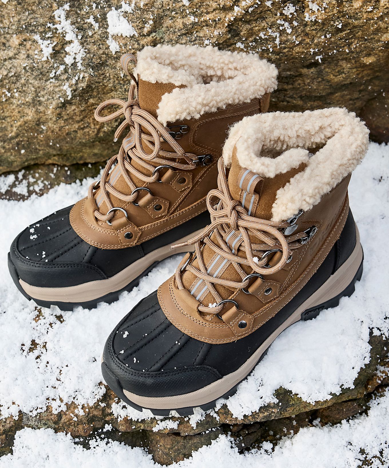 Brune vinterstøvler med svart gummifront, beige pelsfôr og snøring, plassert på snødekket steingrunn.