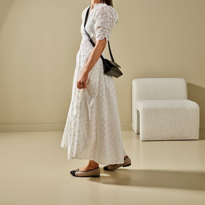 Kvinne iført hvit lang kjole og beige ballerina-sko med svarte tupper, bærer svart skulderveske, står på beige gulv med kremfarget stol i bakgrunnen.