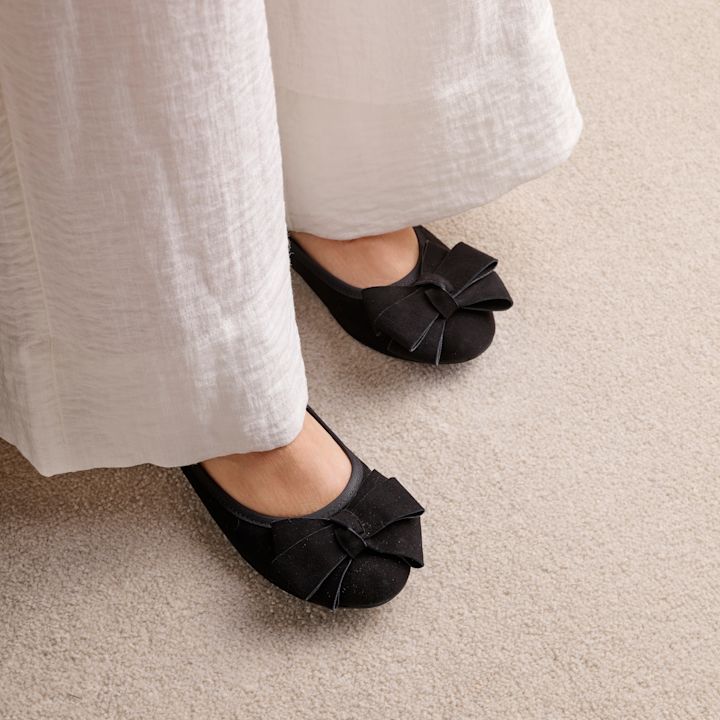 Svarte ballerinasko med sløyfedetalj under hvit linbukse, stående på beige teppe.