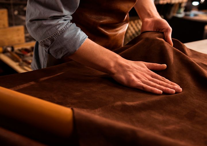 Händer som rör vid och jämnar ut ett stycke brunt läder på ett arbetsbord i en verkstad.