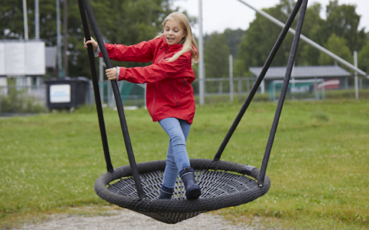 Flicka i röd jacka och blå jeans som leker på en stor rund gunga i en grön park.