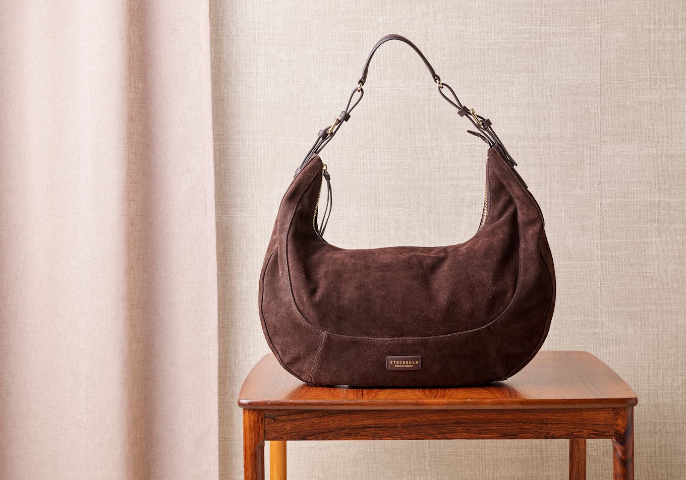 Elegant mörkbrun mockaväska från Stockholm Design Group på ett träbord mot beige bakgrund.
