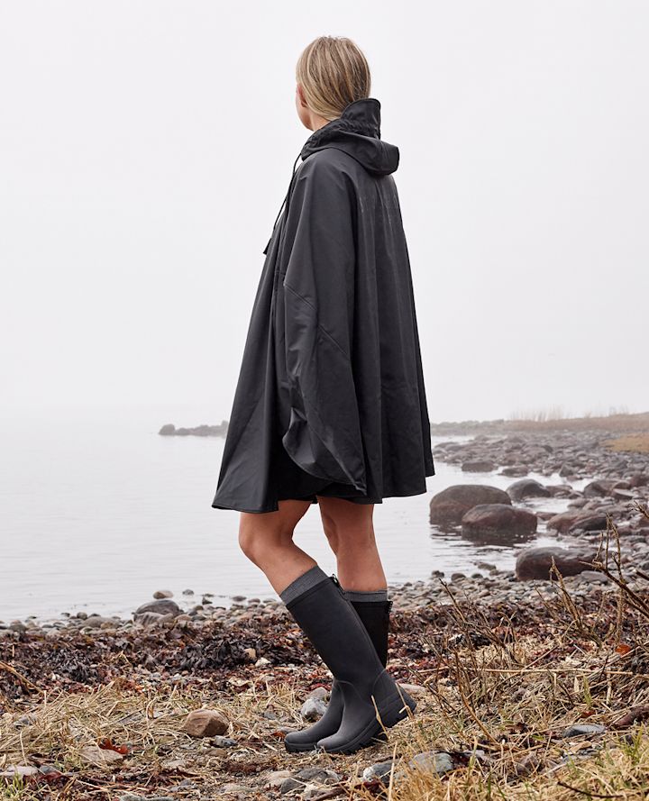 Person står ved en steinete strand iført svart regnponcho og gummistøvler, ser ut mot tåke over vannet.