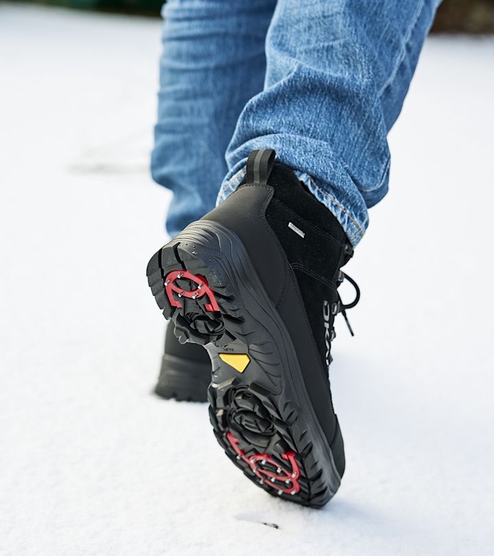 Svart vinterstövel med röda och gula dubbar under sulan, som trampar på snö, iklädd blå jeans.