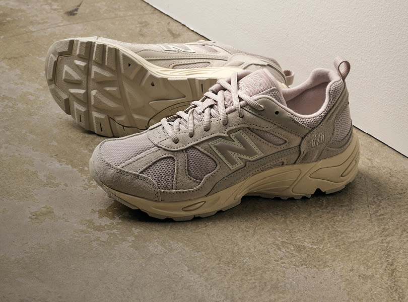 Et par beige New Balance joggesko i sporty stil med mesh-overflate og kraftig sålemønster, plassert på betonggulv.