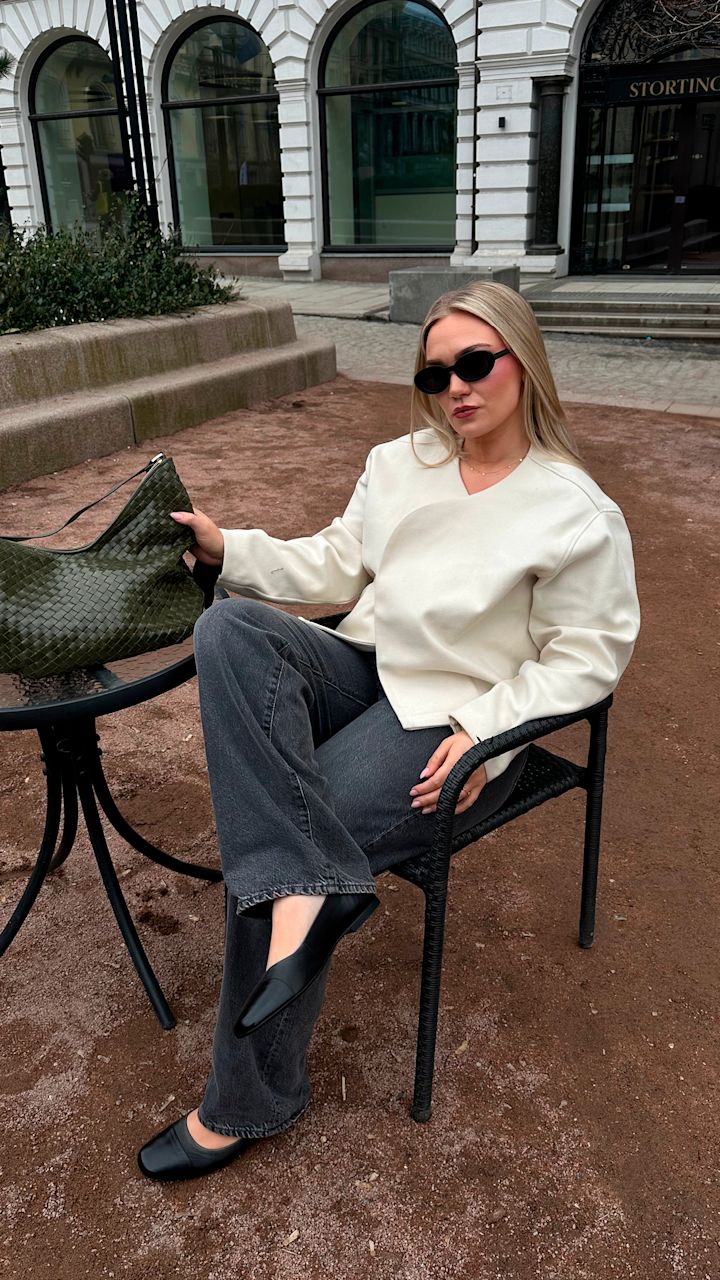 Kvinna med solglasögon sitter i en svart korgstol utomhus, iklädd en vit jacka, mörka jeans och svarta skor, med en grön väska på ett glasbord bredvid.