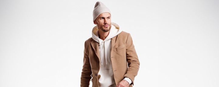 Mann iført beige lue, hvit hettegenser og brun kåpe som ser til siden mot høyre på hvit bakgrunn.