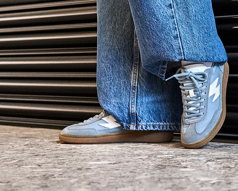 Lyseblå retro sneakers fra Unified med hvitt lynmotiv og gummibunn, brukt med vide jeans foran en metallisk rulleport.