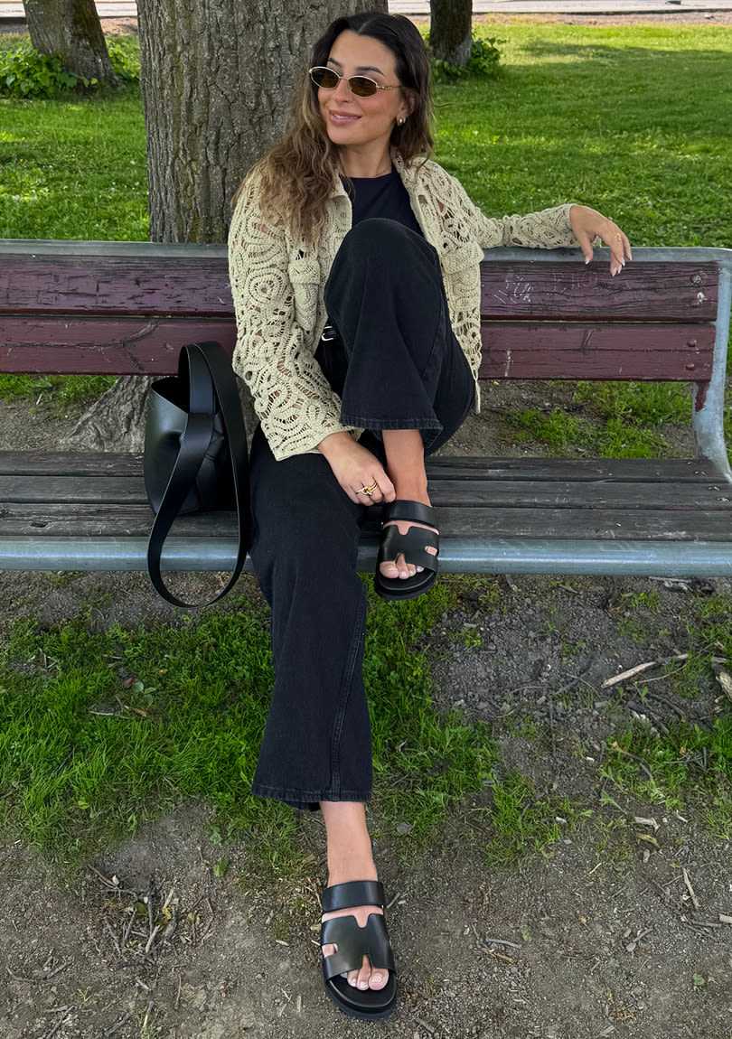 Influenser Sara Boudaya på parkbenk med kremfarget heklet jakke og svart antrekk, iført svarte og trendy skinnsandaler med remmer.