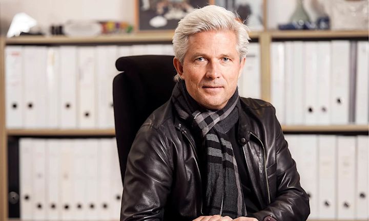 Mann med grått hår iført svart skinnjakke og rutete skjerf sitter i kontorstol foran hylle med hvite permer.