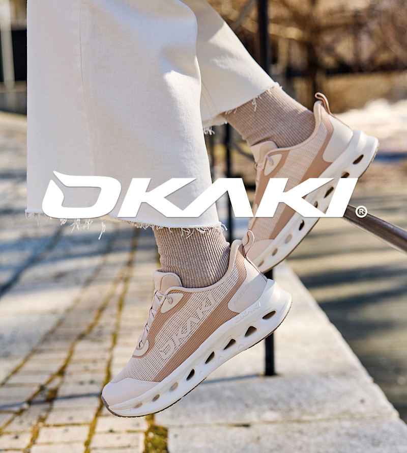 Person med hvite bukser og beige Okaki sportssko med hvit såle og ventilasjonshull, svevende over fortau i sollys.