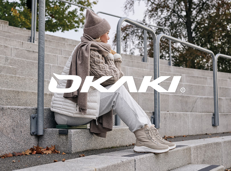 Person i vinterbeklædning sidder på trappe med beige OKAKI støvler i sporty stil og hvid sål. Logo vises i forgrunden.