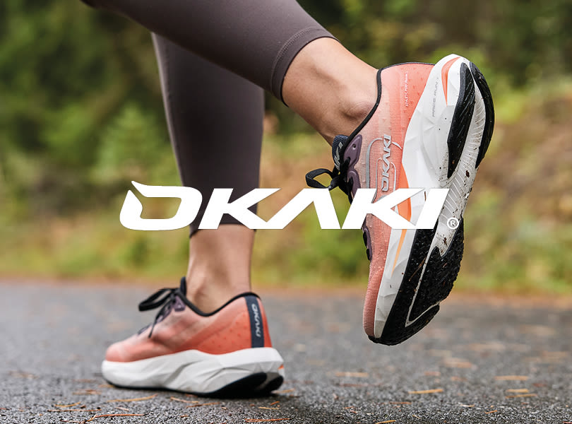 Løper med korallrøde Okaki Pace Run løpesko med hvit såle og svarte detaljer, Okaki-logo vises i forgrunnen.