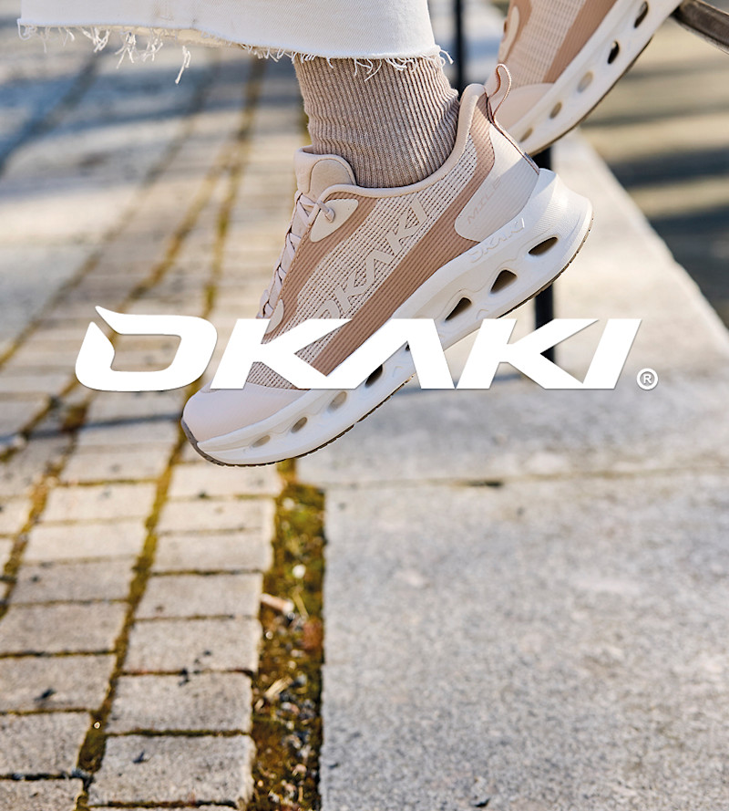 Person med hvite bukser og beige Okaki sportssko med hvit såle og ventilasjonshull, svevende over fortau i sollys.