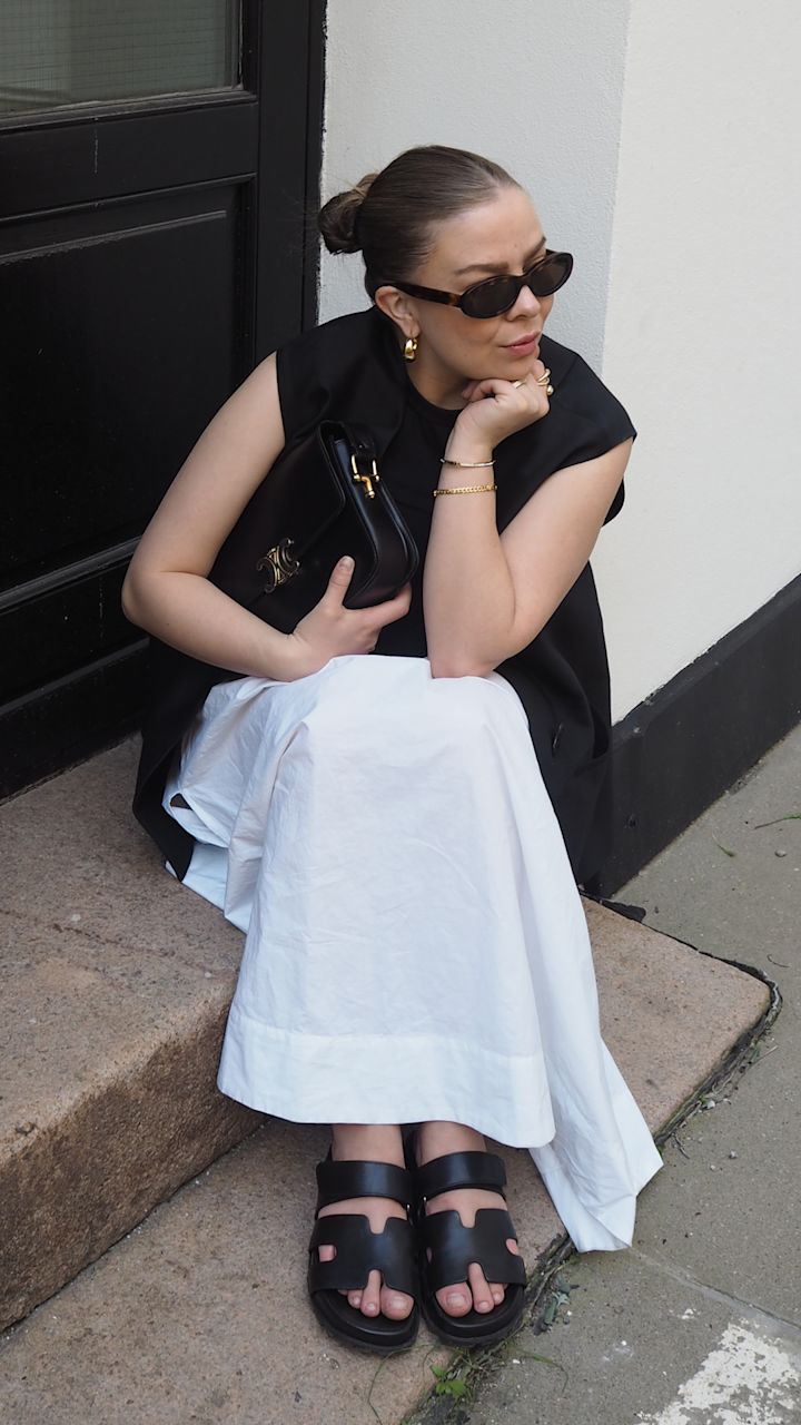 Kvinna med solglasögon och guldsmycken sitter på en trappa utanför en byggnad, klädd i svart topp, vitt långt kjol och svarta sandaler, håller en svart väska.