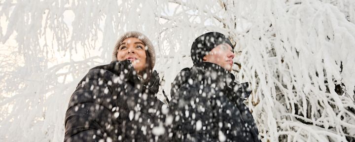 Två personer i varma vinterjackor och mössor står rygg mot rygg medan snö faller framför dem i ett snötäckt skogsområde.