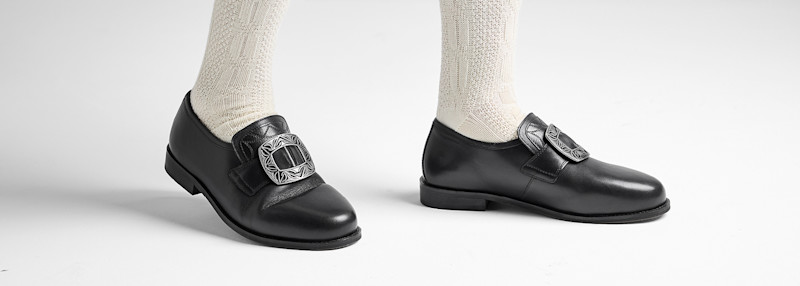 Svart lærsko med stor dekorativ spenne på foten iført hvite strikkede sokker på hvit bakgrunn.