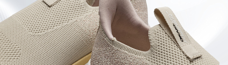 Beige stickade sneakers med textilyta, elastisk häl och ögla bak, visade i närbild mot vit bakgrund.