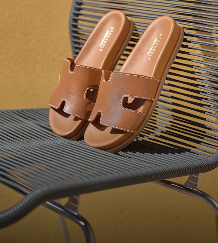 Brune flate sandaler i skinn med bred utskåret rem, plassert på en metallstol med gul bakgrunn.