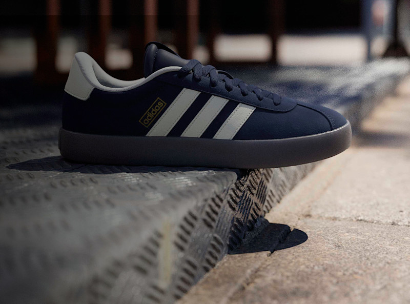 Marinblå Adidas sneaker med vita ränder och grå sula, placerad på en betongkant i urban miljö.