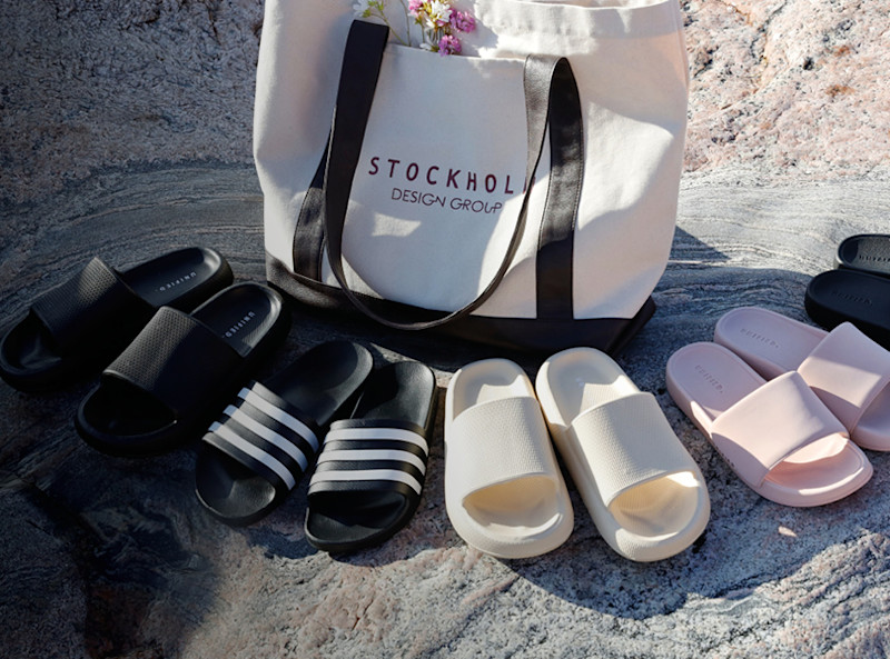 Fem par slippers i svart, hvit og rosa på stein, ved siden av en hvit tote-bag med teksten Stockholm Design Group.