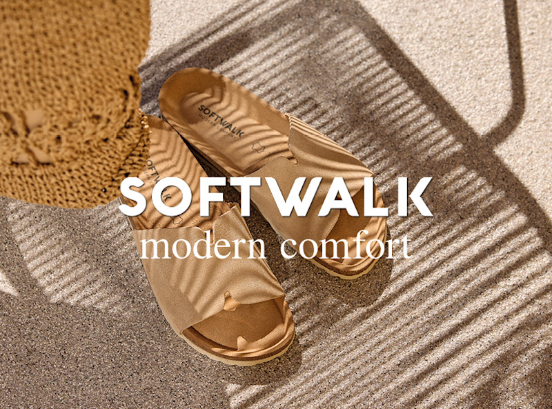 Beige, flate sandaler fra Softwalk med bred reimdetalj, fotografert ovenfra på sandfarget underlag ved siden av en stråhatt.