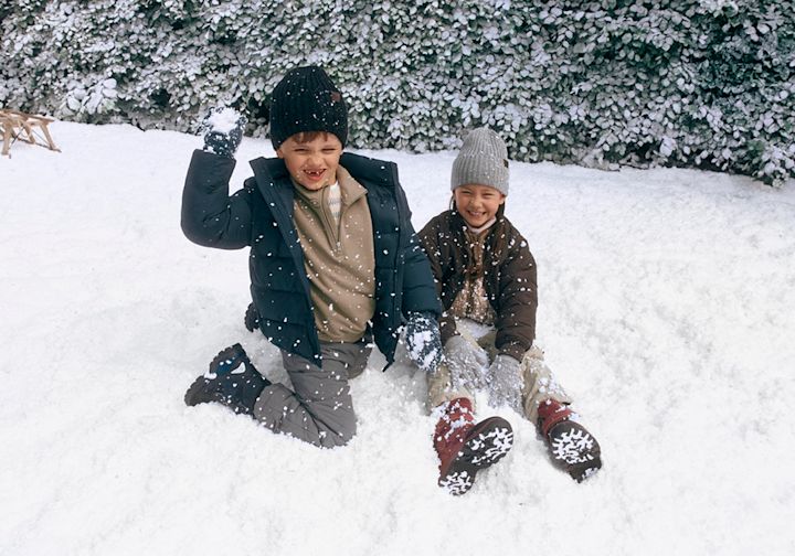 Två barn leker i snön, klädda i vinterkläder. Ett barn har svarta vinterstövlar och det andra har röda snökängor med snöflingemönster.