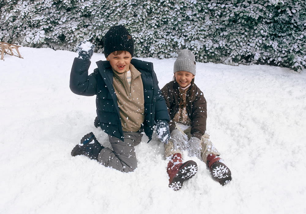 Två barn leker i snön, klädda i vinterkläder. Ett barn har svarta vinterstövlar och det andra har röda snökängor med snöflingemönster.