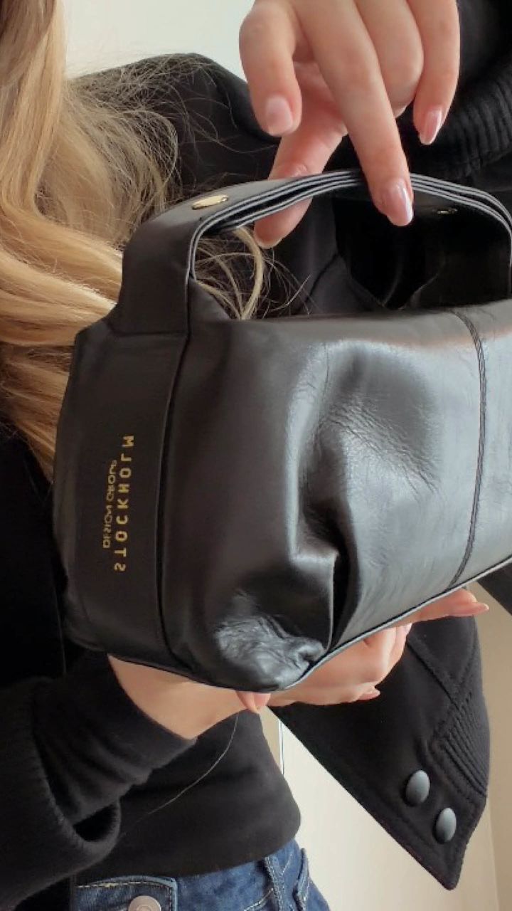 Person håller en svart läderväska med guldtext, klädd i svart jacka och jeans. Blont hår syns i bakgrunden.