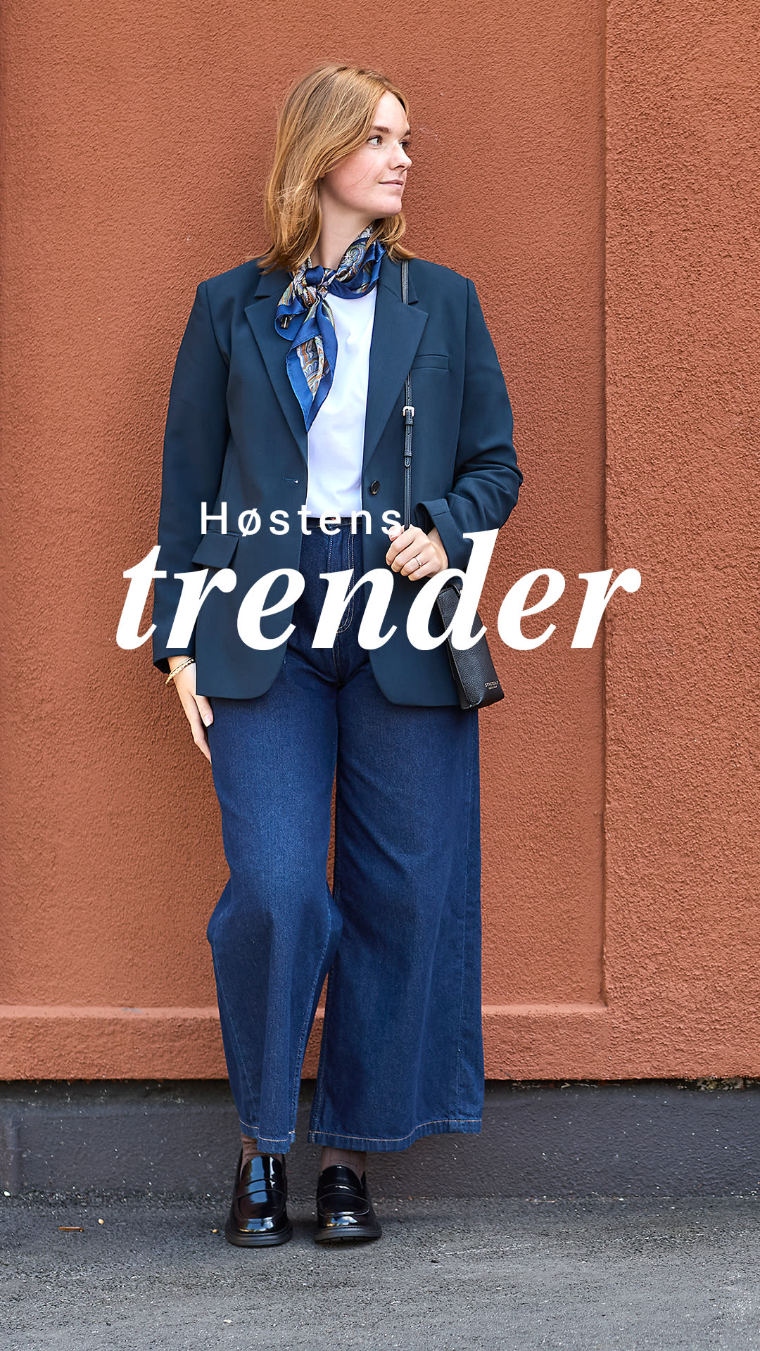 Person i mørkeblå blazer og jeans mot terrakottavegg, med svarte blanke loafers. Tekst: "Høstens trender".