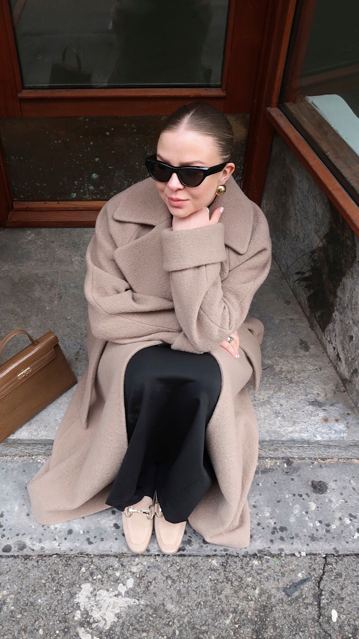 Kvinna med bruna solglasögon, beige kappa, svart kjol och beige skor sitter på trottoarkanten vid entrédörren med en brun handväska bredvid sig.