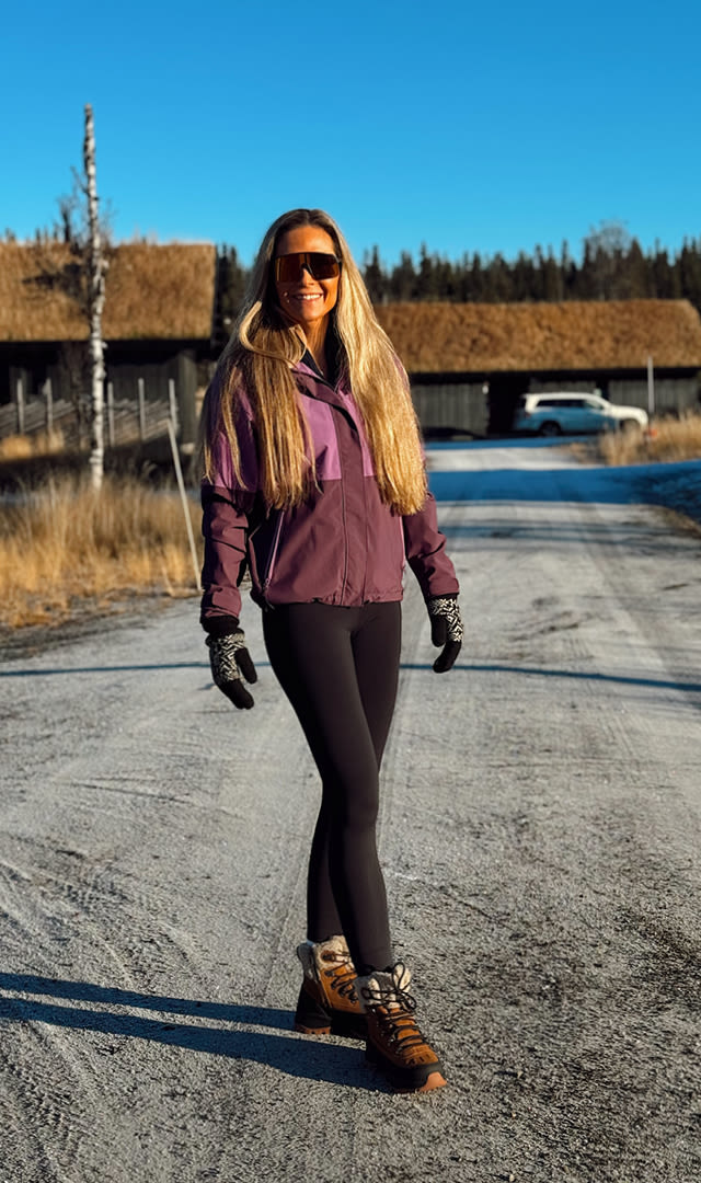 Kvinna med långt ljust hår går på grusväg iklädd lila jacka, svarta tights, bruna vandringsskor, vantar och solglasögon, med stugor och skog i bakgrunden.