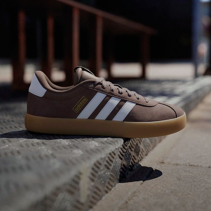 Adidas sneaker i brun semsket skinn med hvite striper og gummisåle, plassert på en metalloverflate utendørs.