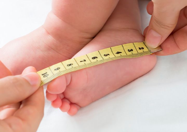 Babyfot mäts med ett måttband hålls av två händer mot en vit bakgrund.