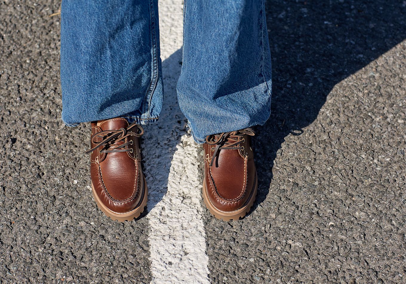 Brune skinnsko i båtstil med snøring og kraftig såle, brukt med blå jeans, stående på asfalt med hvit stripe.