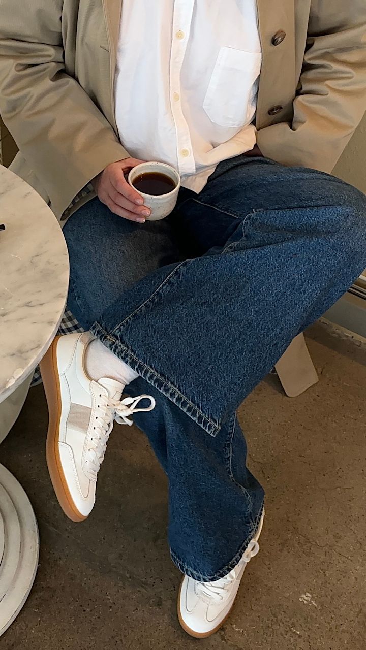 Person sitter med bena krysset iført blå jeans, hvite sneakers og beige jakke, holder en kopp kaffe ved et marmorbord.