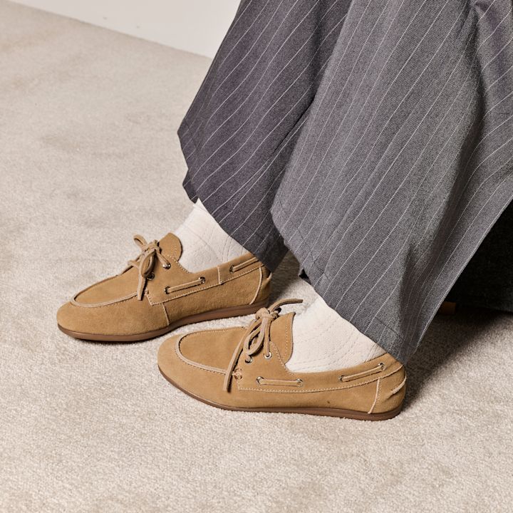Lysebrune semsket båtsko med snøring, brukt med hvite sokker og grå stripete bukser på et beige teppe.