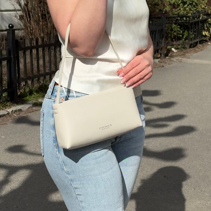 Person i hvit topp og lyseblå jeans holder en kremfarget veske fra Stockholm Design Group på en solrik dag utendørs.