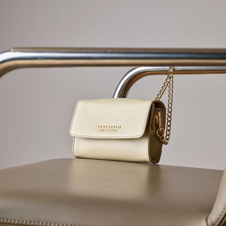 Liten beige veske med gullkjede henger på ryggstøtten til en stol med metallramme og brunt sete.