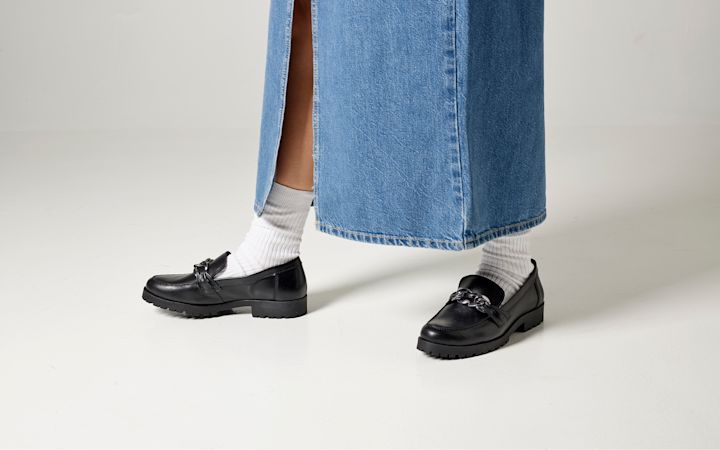 Sorte skinnsko med tykk såle og kjededetalj på føttene til en person iført hvite sokker og en lang blå denimskjørt med splitt.