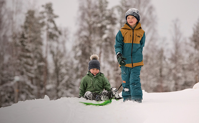 Två barn i vinterkläder leker i snön, ett sitter på en grön pulka medan det andra står bredvid i blå vinterstövlar.