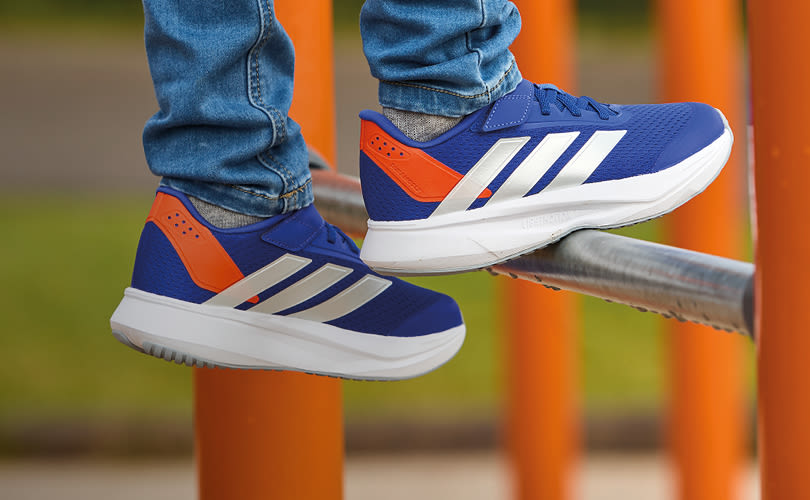Blå og oransje Adidas sneakers med hvite striper, brukt med jeans, balanserer på en metallstang i en lekeplass.
