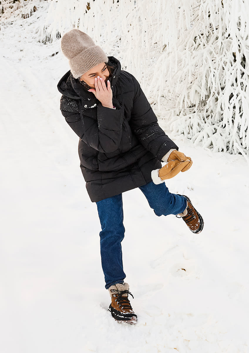 Person i vinterklær med beige lue og brune vinterstøvler med pelskant leker i snøen omgitt av snødekte trær.